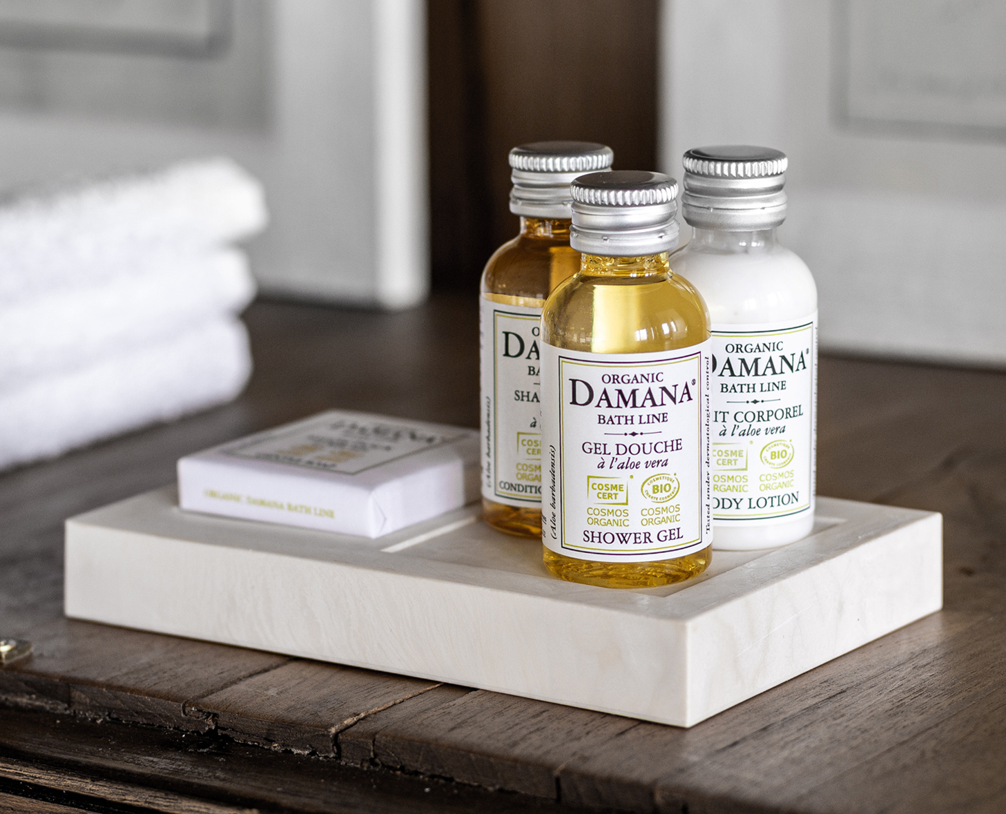 Três produtos de banho ecológicos — gel de banho, loção corporal e champô — em pequenos frascos de vidro, expostos numa bandeja de madeira clara, ao lado de um sabonete e toalhas brancas cuidadosamente arrumadas.