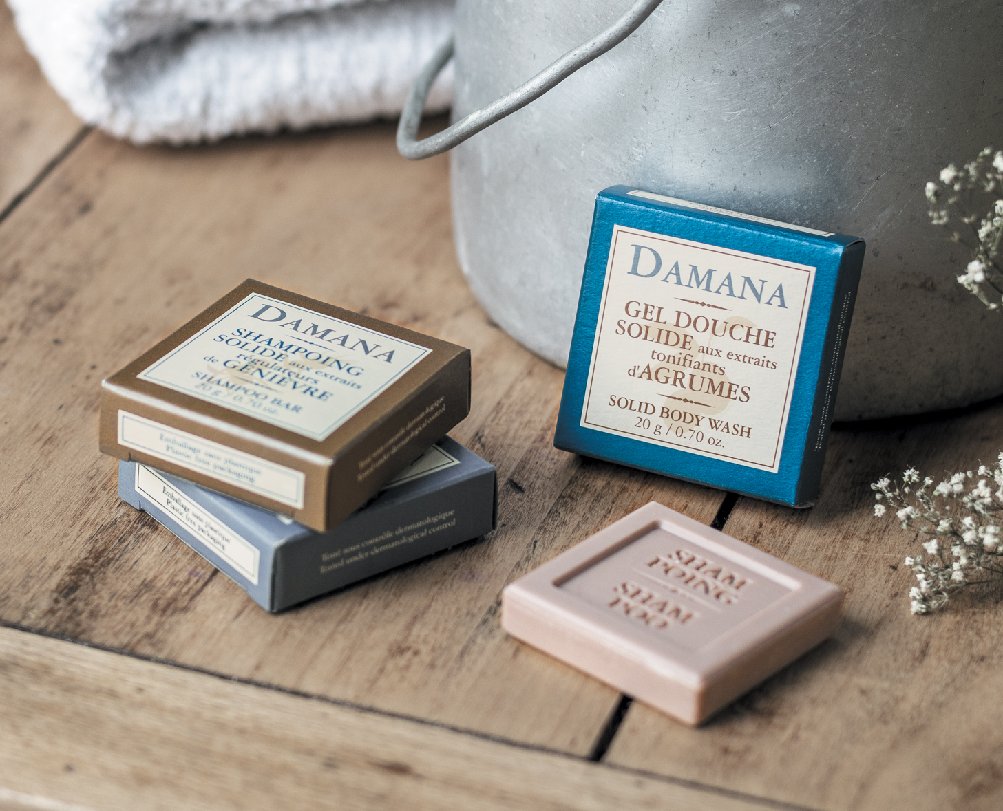 Una collezione di prodotti da bagno solidi colorati, tra cui shampoo e bagnoschiuma, disposti accanto a un secchio di metallo su una superficie di legno. Sullo sfondo è visibile anche un piccolo bouquet di fiori secchi.