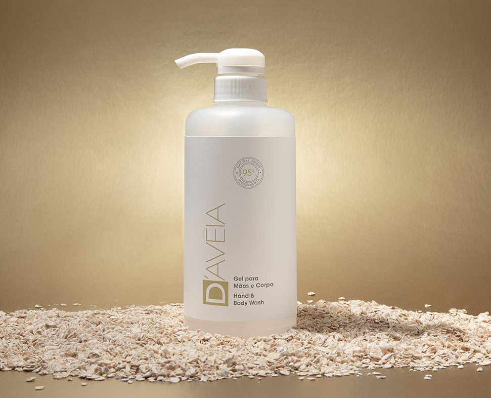 Um frasco transparente de sabonete líquido para mãos e corpo repousa sobre uma cama de aveia, com um fundo dourado suave destacando o produto. O rótulo apresenta o nome da marca «DAVEIA» em fonte elegante.