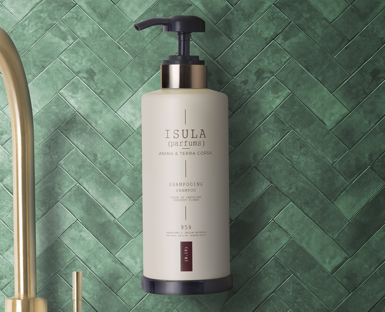Una bottiglia di shampoo beige con un erogatore nero è appesa su una parete rivestita di piastrelle verdi a spina di pesce, con l'etichetta "ISULA parfums)" e altre informazioni sul prodotto. Sullo sfondo è parzialmente visibile un rubinetto dorato.