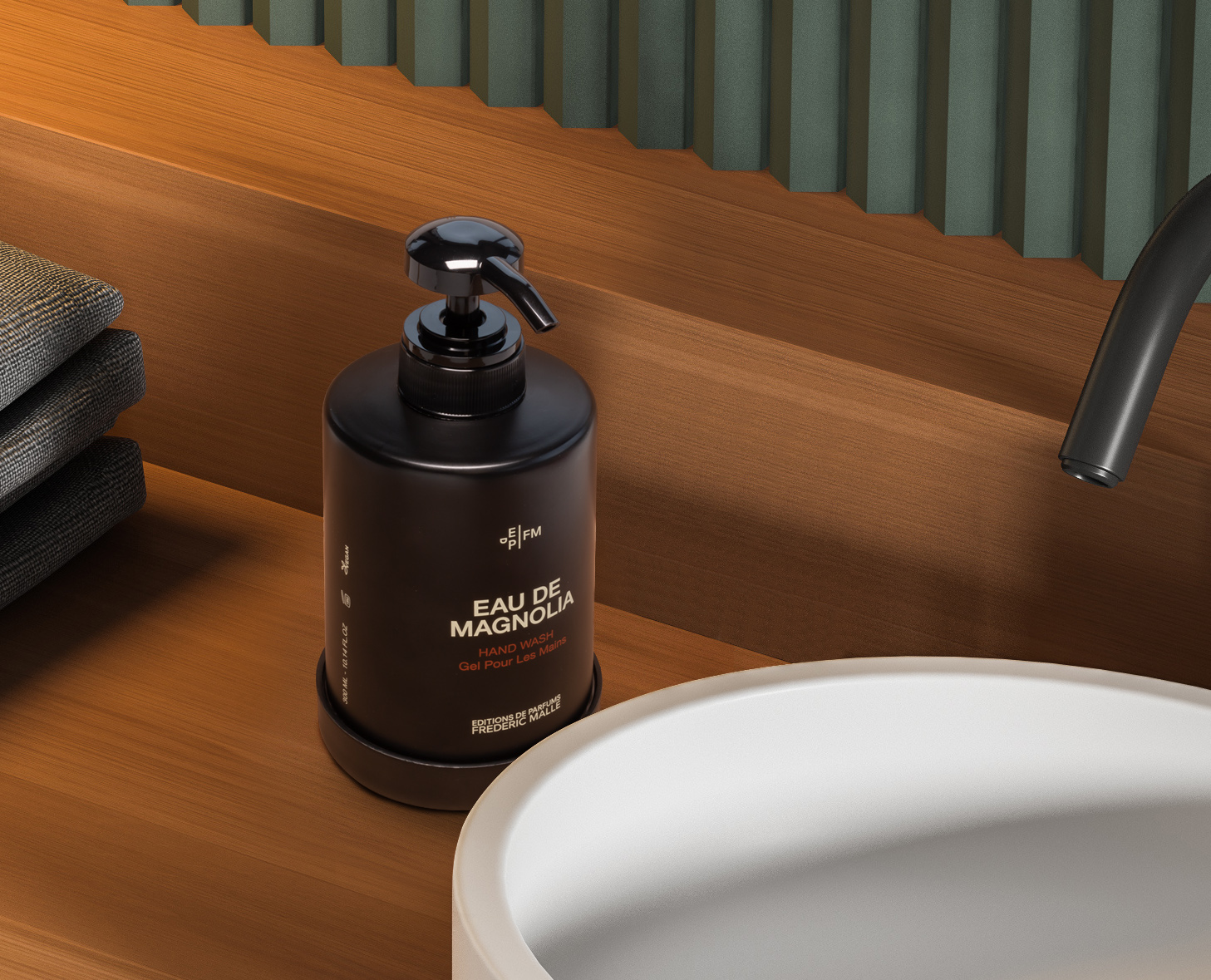 Un elegante flacone scuro di sapone liquido per le mani "Eau de Magnolia" è posizionato su un ripiano in legno accanto a asciugamani grigi ordinatamente impilati e un lavandino moderno. Lo sfondo è caratterizzato da pannelli verdi testurizzati.