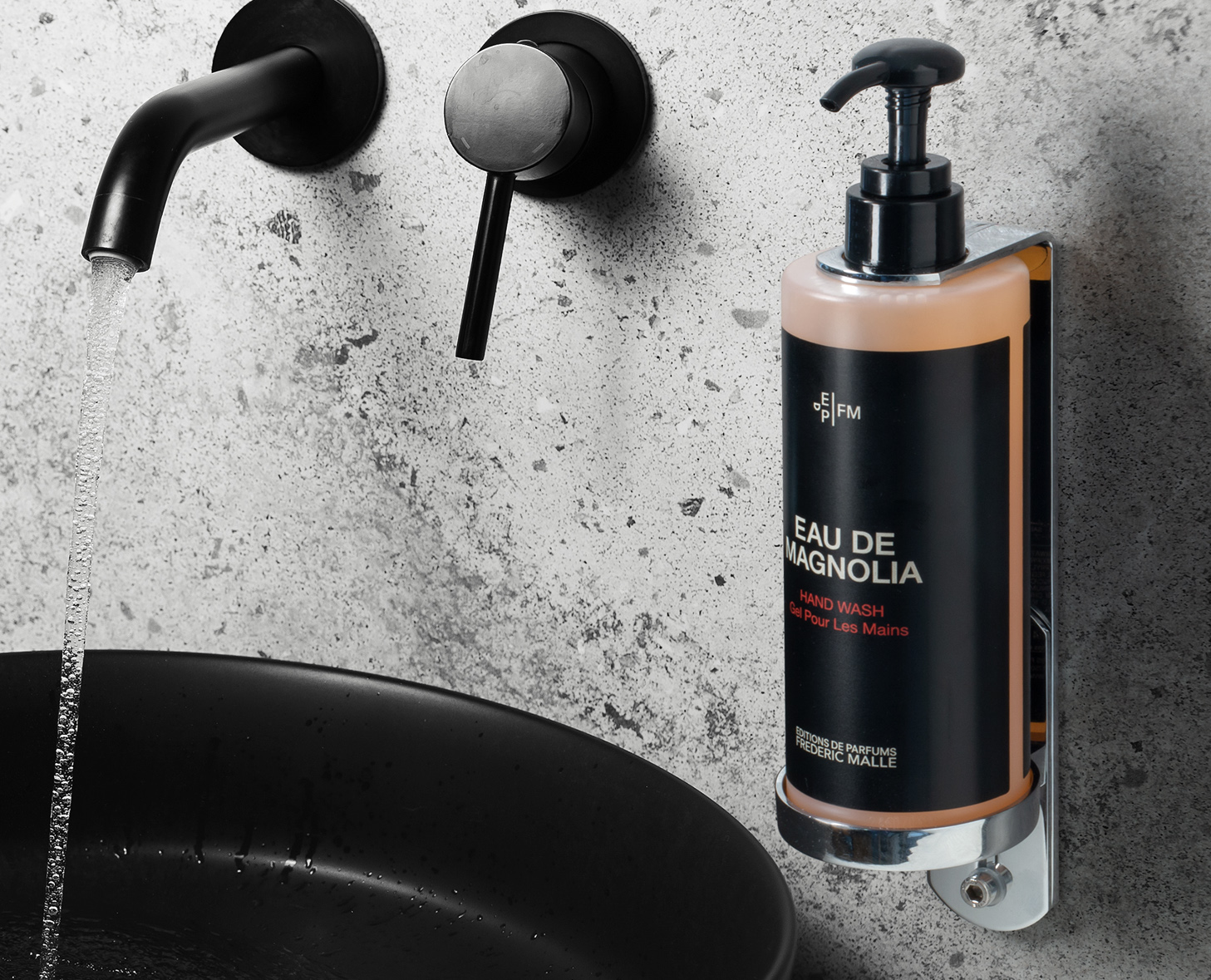 Un dispenser di sapone liquido nero e oro con l'etichetta "Eau de Magnolia" montato su una parete accanto a un elegante lavabo nero e un rubinetto moderno. L'acqua scorre dal rubinetto nel lavabo.