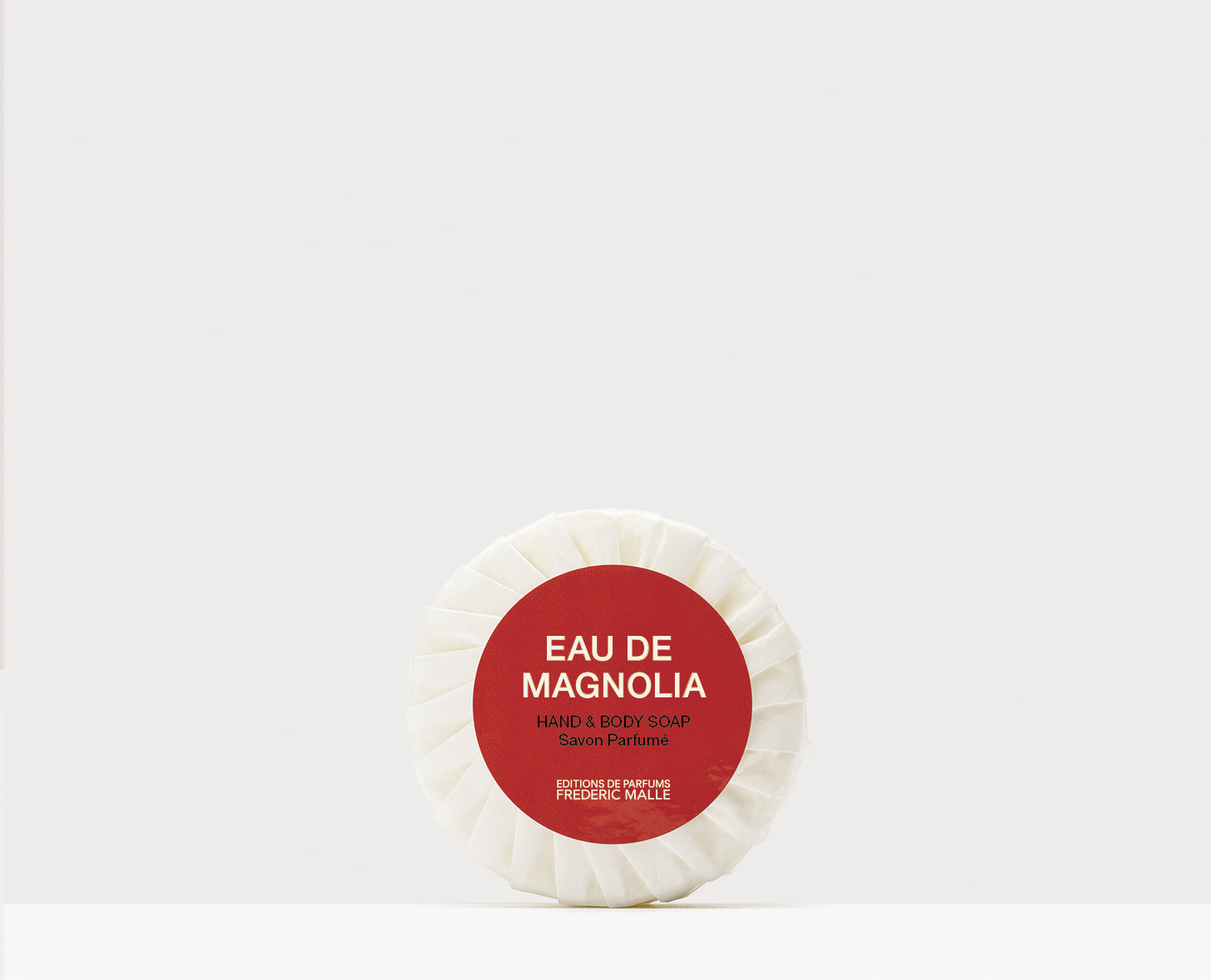 Una saponetta rotonda con la scritta "Eau de Magnolia" e un disegno rosso e bianco, posizionata su uno sfondo bianco uniforme.