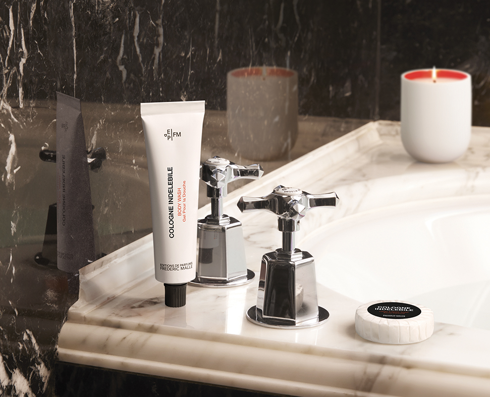 Eine elegante Badezimmer-Arbeitsplatte mit einer Tube Kölnischwasser, Wasserhahngriffen im Vintage-Stil, einem kleinen Glas und einer Kerze, alles vor einem Marmorhintergrund.