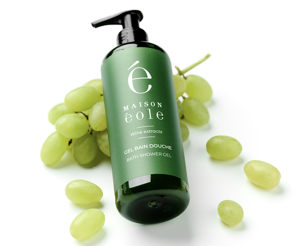 Una bottiglia verde di gel doccia con pompa, con l'etichetta "MAISON éole" e illustrazioni di uva, circondata da grappoli d'uva verde.