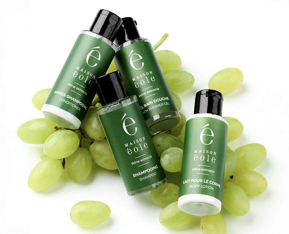 Quattro flaconi di colore verde contenenti prodotti per la cura dei capelli e del corpo sono disposti tra grappoli d'uva fresca. Le etichette riportano il nome del marchio "MAISON ÉOLE" e sono principalmente in francese.