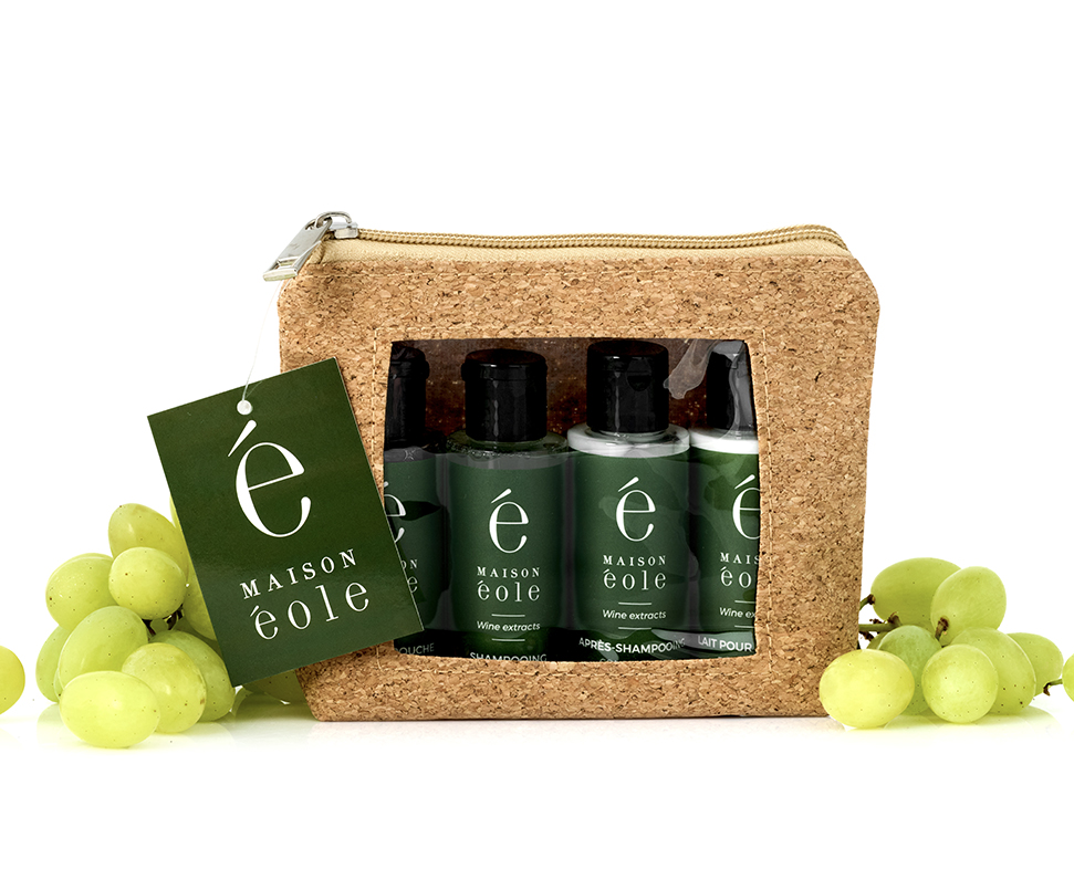 Una pochette in sughero con chiusura a zip contenente prodotti per la cura dei capelli e del corpo in formato da viaggio in flaconi verdi, accompagnati da freschi acini d'uva verde.