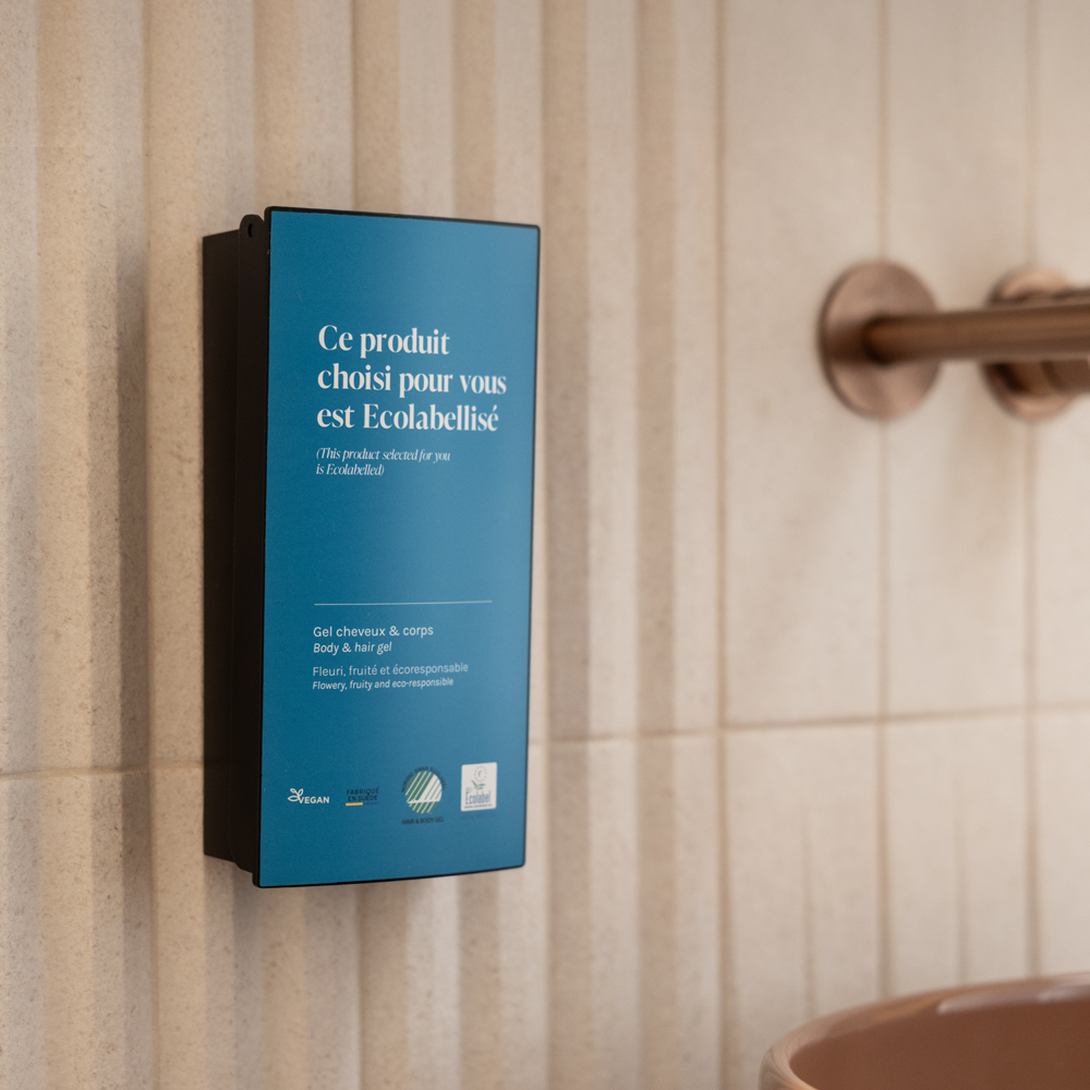 Een wanddispenser met milieuvriendelijke haar- en lichaamsgel, voorzien van een blauw etiket met Franstalige tekst die aangeeft dat het product Ecolabellisé is.