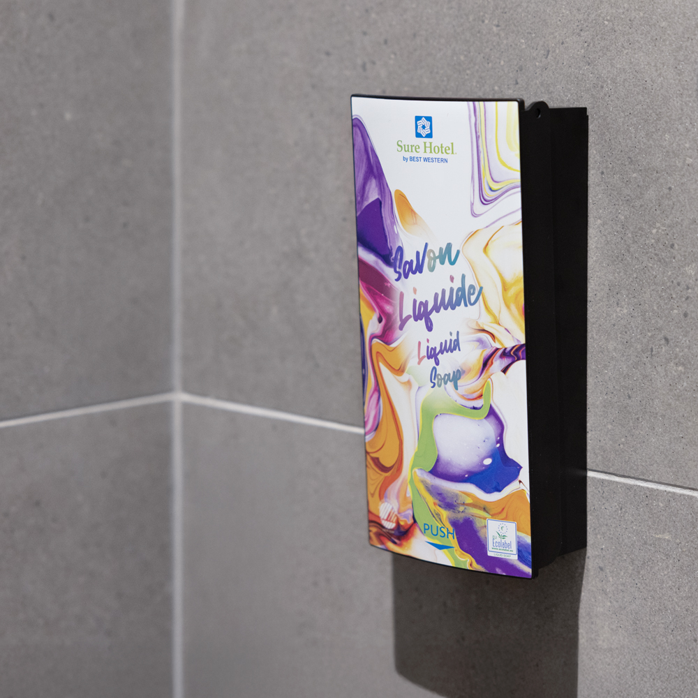 Een wandgemonteerde vloeibare zeepdispenser met kleurrijke, abstracte verpakking in een moderne badkamer. Op de dispenser staat "Sure Hotel" en "Liquid Soap" (vloeibare zeep).