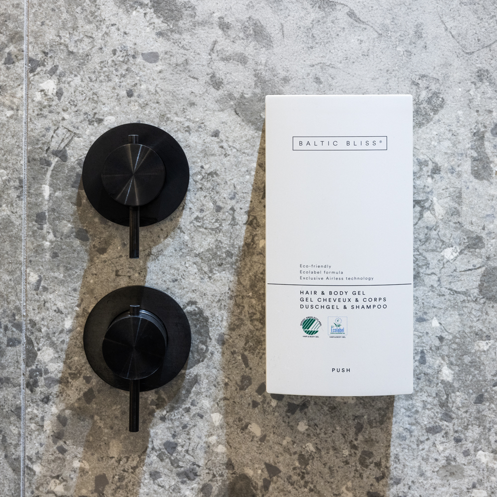 Een strakke witte doos met het opschriftBALTIC BLISS' en een milieuvriendelijk logo, geplaatst naast twee zwarte, aan de muur bevestigde dispensers voor shampoo en douchegel op een grijs oppervlak met structuur.