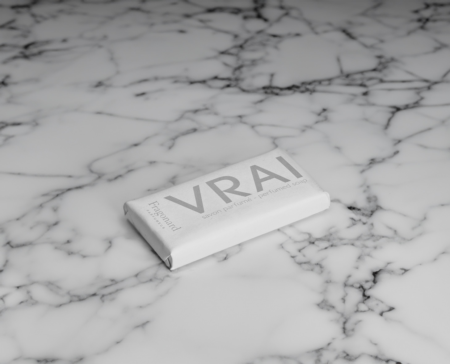 Una saponetta avvolta in carta bianca minimalista con la parola "VRAI" stampata in grassetto, appoggiata su un ripiano in marmo.