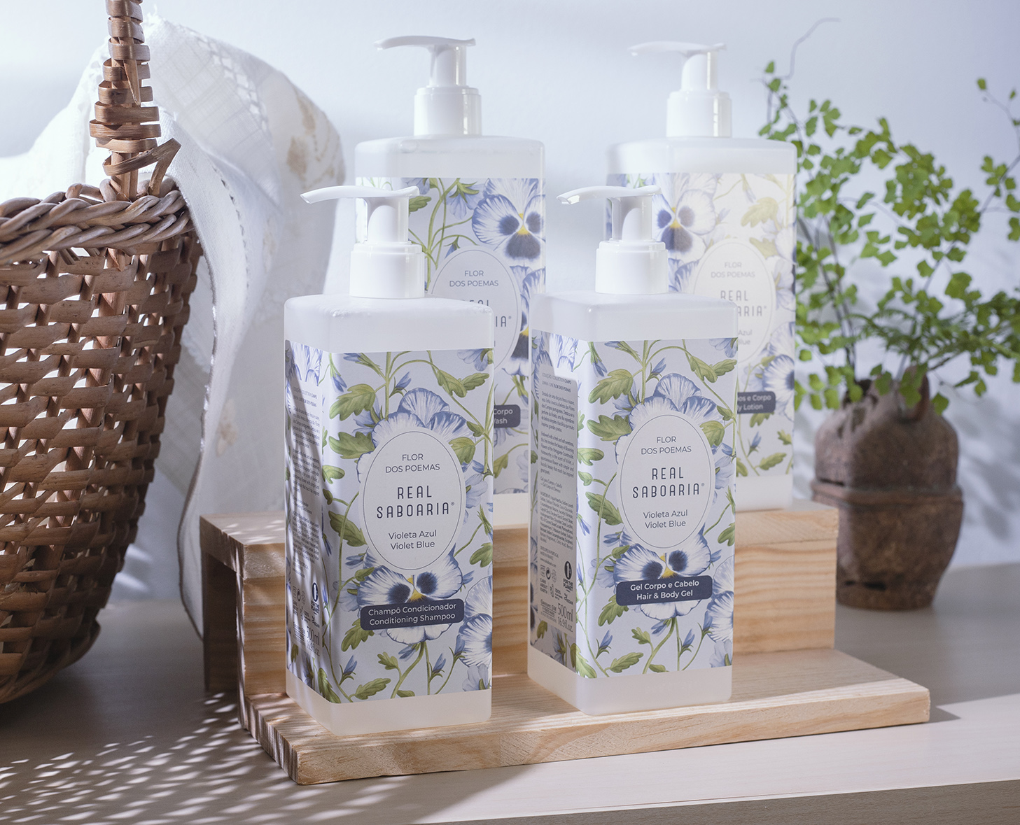 Quattro bottiglie di sapone liquido disposte ordinatamente su un supporto di legno, con motivi floreali ed etichette. Sullo sfondo, un cesto intrecciato e una piccola pianta decorativa.