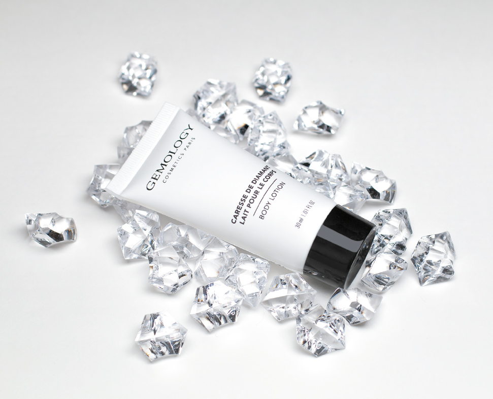 Eine Tube Gemology creme ist von verstreuten klaren Kristallsteinen vor einem schlichten weißen Hintergrund umgeben.