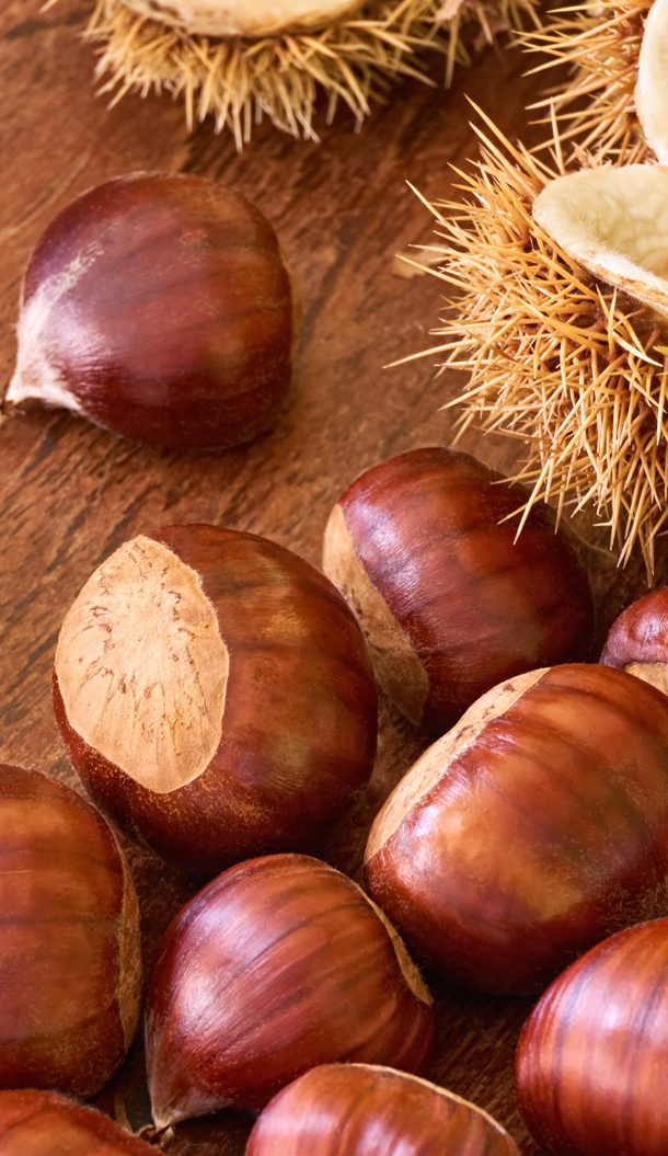 Primo piano di castagne marroni lucide sparse su una superficie di legno, con un guscio parzialmente aperto che mostra la buccia esterna appuntita.