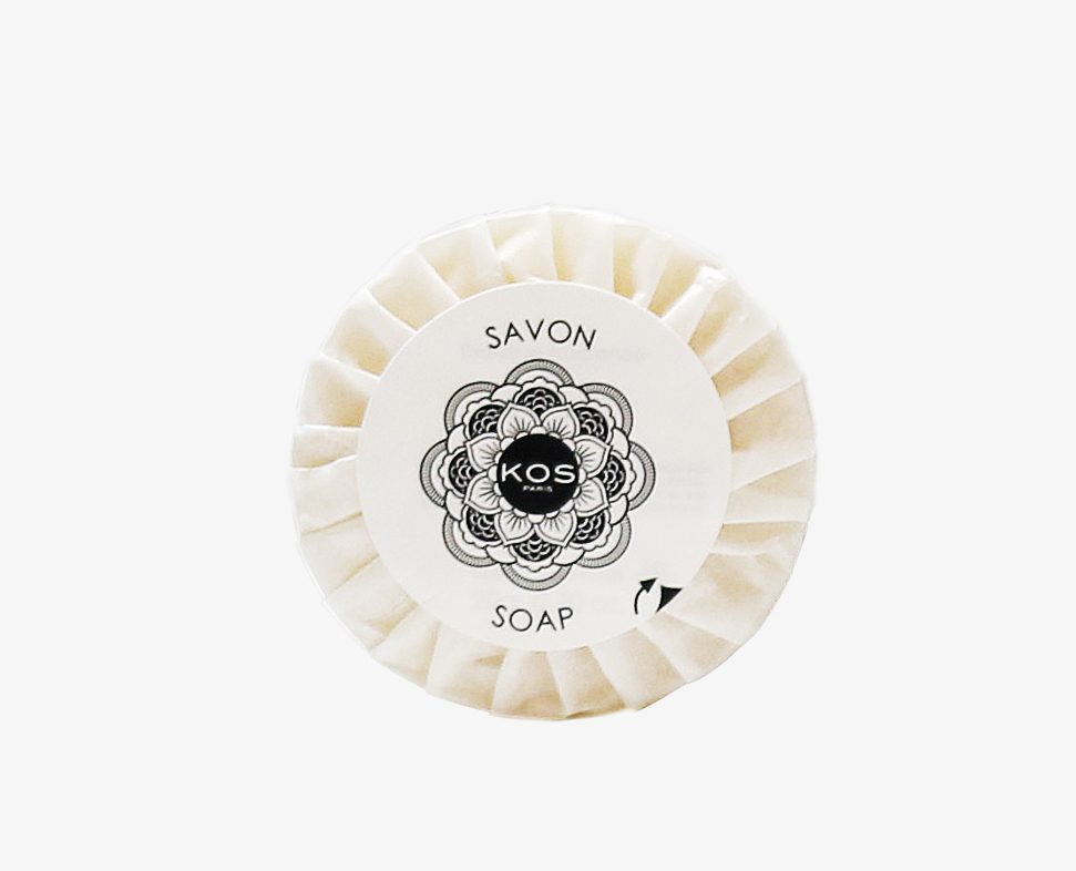 Uma barra de sabão redonda com um exterior padronizado e uma etiqueta com a inscriçãoKOS Soap» em letras pretas. O fundo é branco liso.
