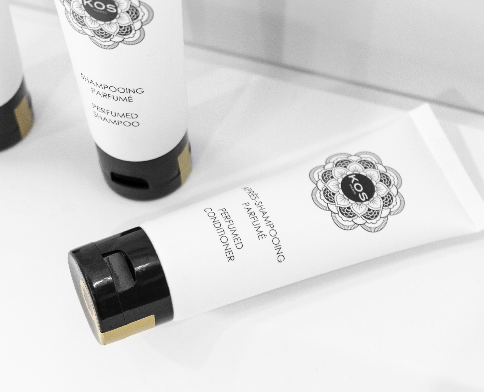 Uma bisnaga de condicionador perfumado com um rótulo decorativo preto e branco, ao lado de dois frascos semelhantes de champô. As bisnagas apresentam um desenho floral e o nome da marca «KOS».