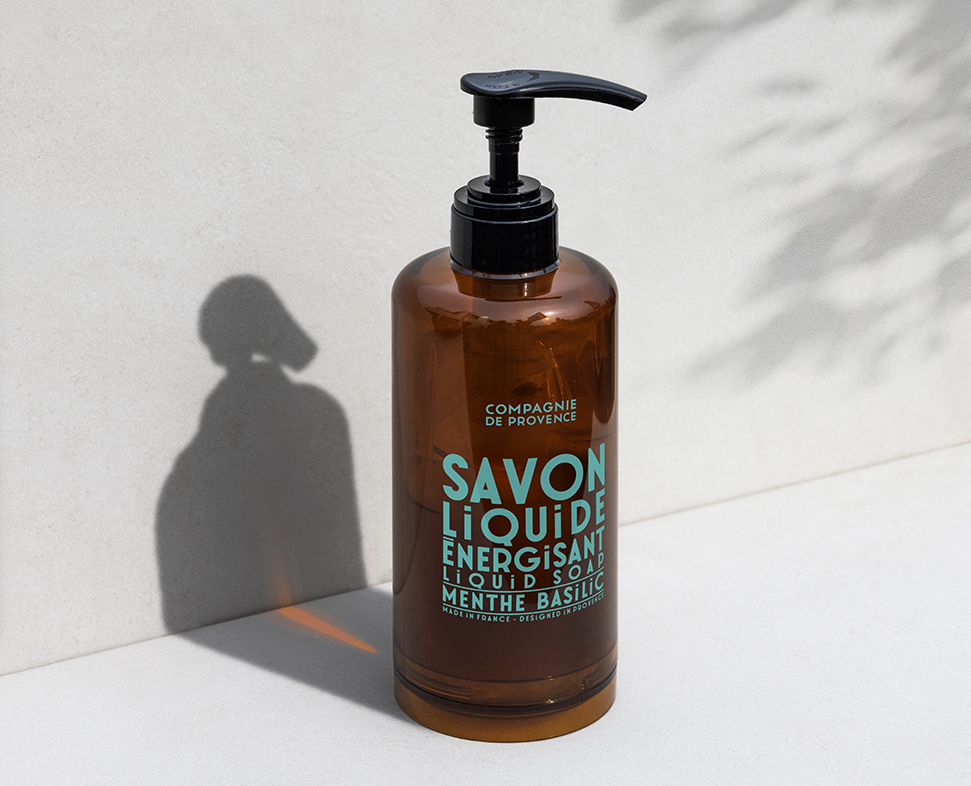 Una bottiglia marrone con pompa etichettata "Savon Liquide Energisant Menthe Basilic" è appoggiata su una superficie chiara, proiettando un'ombra.