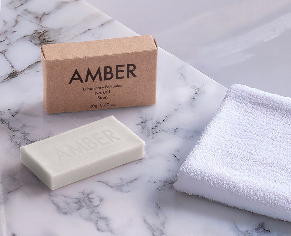 Una saponetta profumata all'ambra con la scritta "AMBER" accanto a un asciugamano bianco, su una superficie di marmo. La saponetta è contenuta in una semplice scatola di cartone marrone.