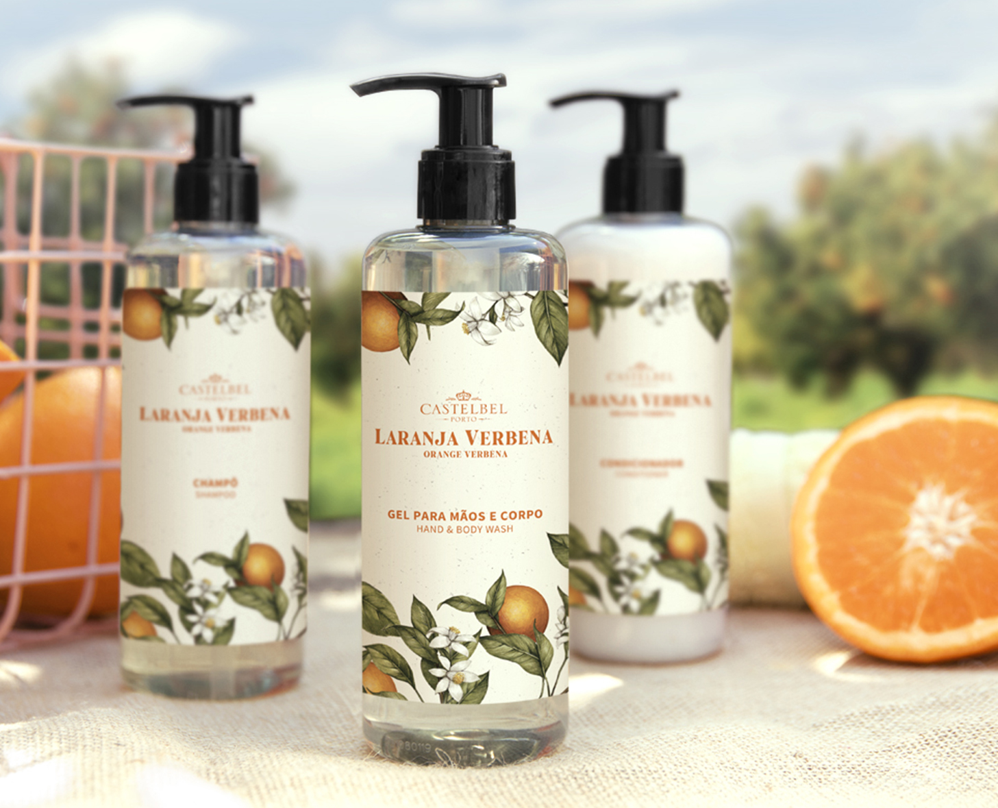 Una collezione di tre flaconi con l'etichetta "Laranja Verbena" contenenti un detergente per mani e corpo, uno shampoo e un balsamo, sullo sfondo di un paesaggio naturale con arance.