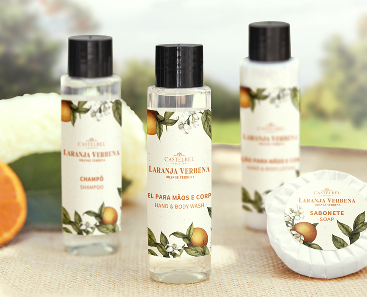 Tre flaconi di cosmetici e una saponetta con l'etichetta "Castelbel Laranja Verbena" con motivi floreali e arancioni, disposti su uno sfondo chiaro con un tocco di verde.