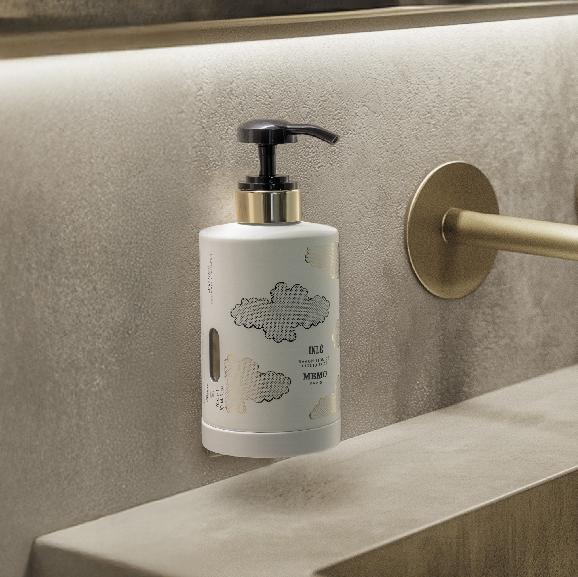 Un flacone bianco con pompa, decorato con un motivo a nuvole e dettagli dorati, è appoggiato su una parete in pietra grezza, accanto a un rubinetto dorato in un bagno moderno.