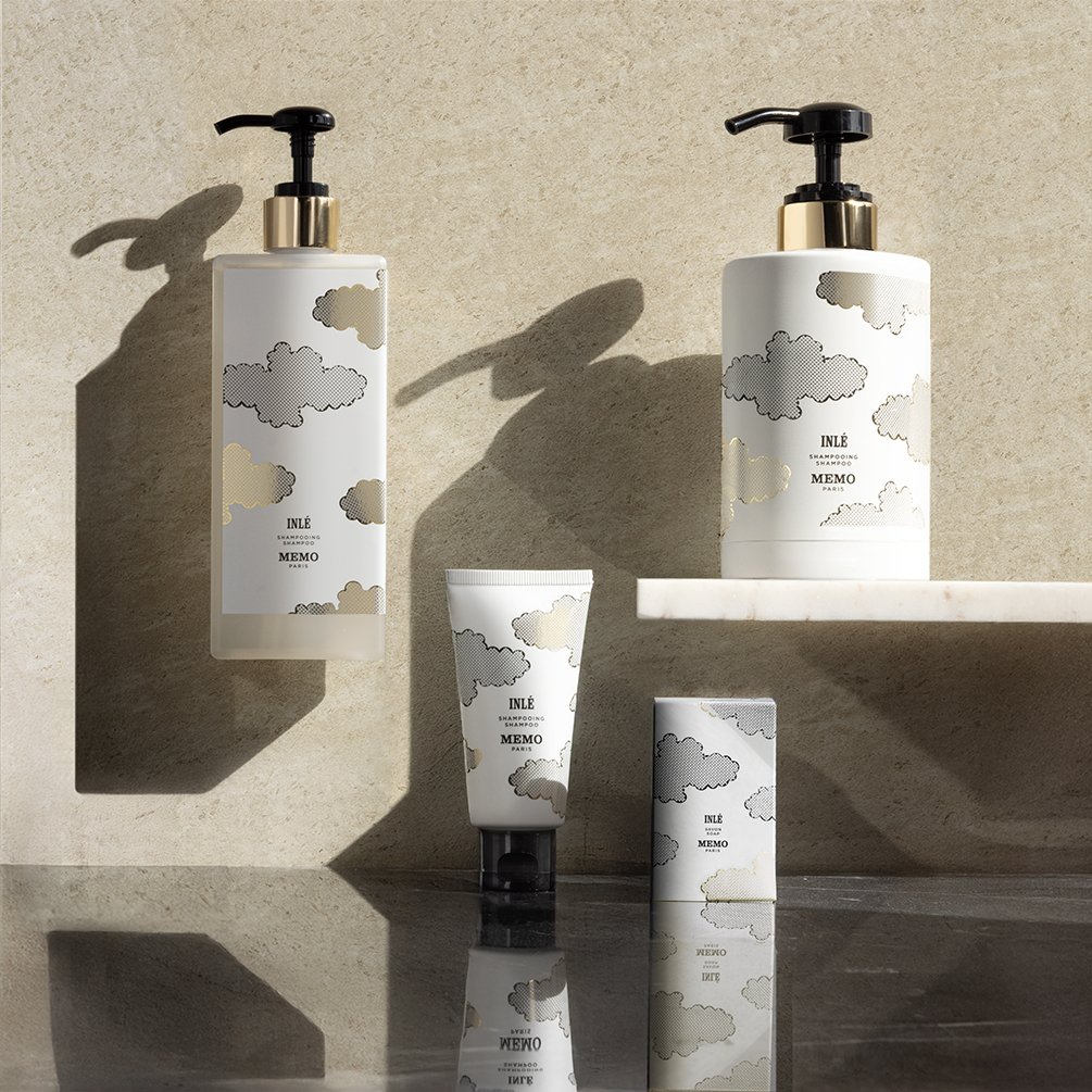 Tre eleganti prodotti per la cura del corpo sono esposti su uno sfondo beige testurizzato, caratterizzati da un lussuoso packaging bianco e oro con motivi a nuvole.