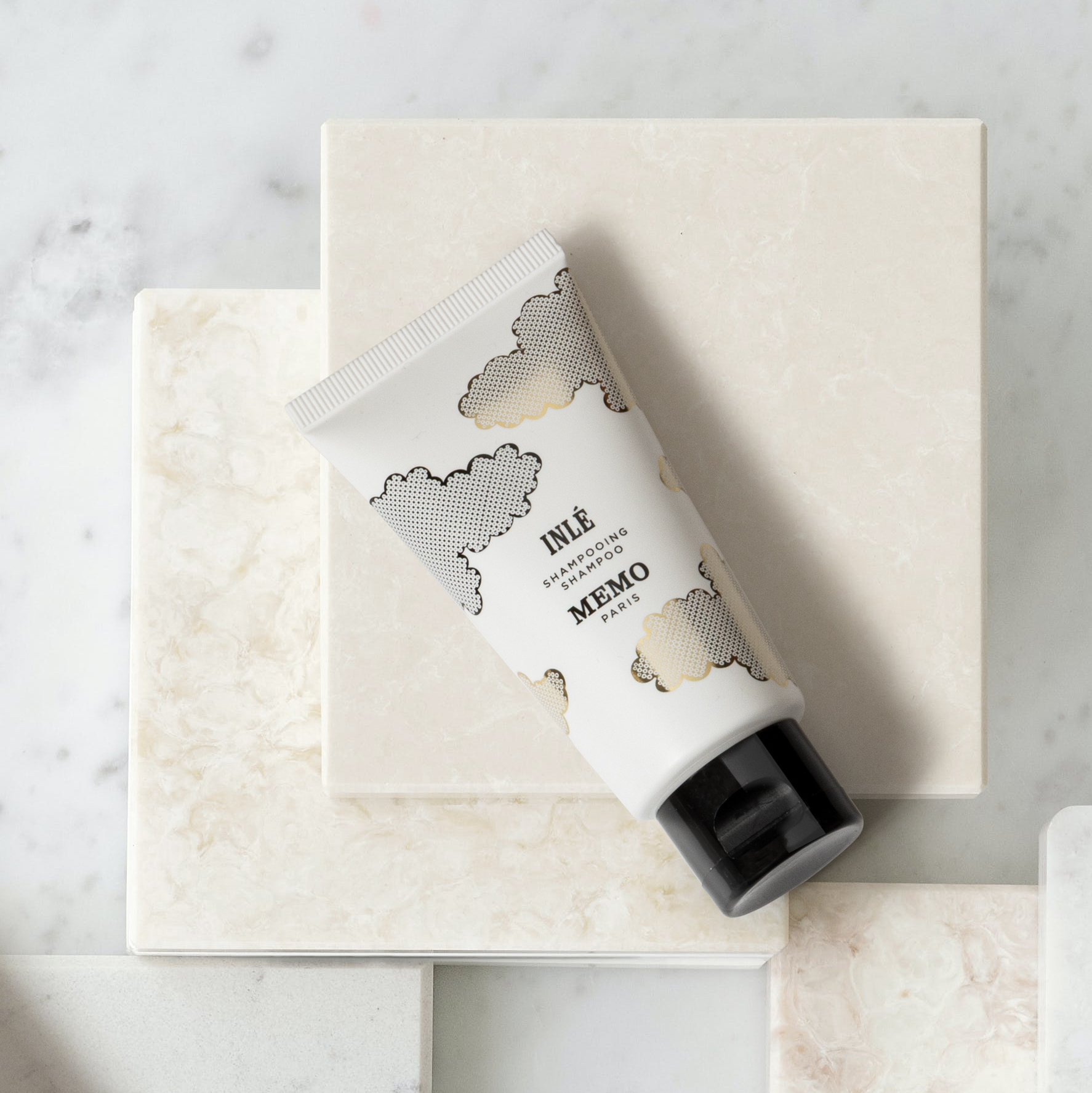 Un tubetto di shampoo con disegni di nuvole è appoggiato su una superficie di marmo accanto a piastrelle di marmo impilate. La confezione è bianca con dettagli neri.