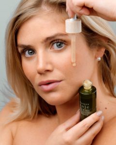 Una donna con i capelli biondi sta applicando una goccia di siero per il viso sulla pelle, tenendo una bottiglia verde nell'altra mano. Ha un'espressione neutra e guarda verso la telecamera.