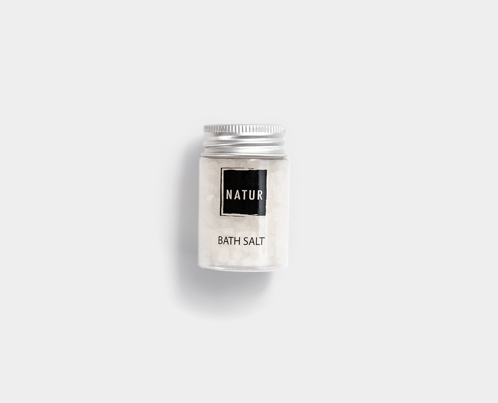 Un piccolo barattolo trasparente di sali da bagno con coperchio argentato, con un'etichetta nera su cui è scritto "NATUR SALT".