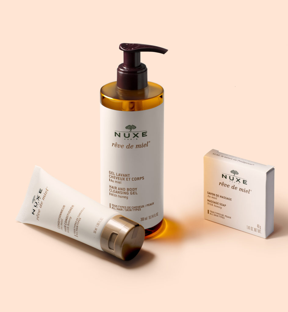 Um frasco de gel de limpeza para cabelo e corpo NUXE Rêve de Miel, um tubo de hidratante NUXE e uma pequena caixa de sabonete, todos dispostos sobre um fundo suave em tom pêssego.