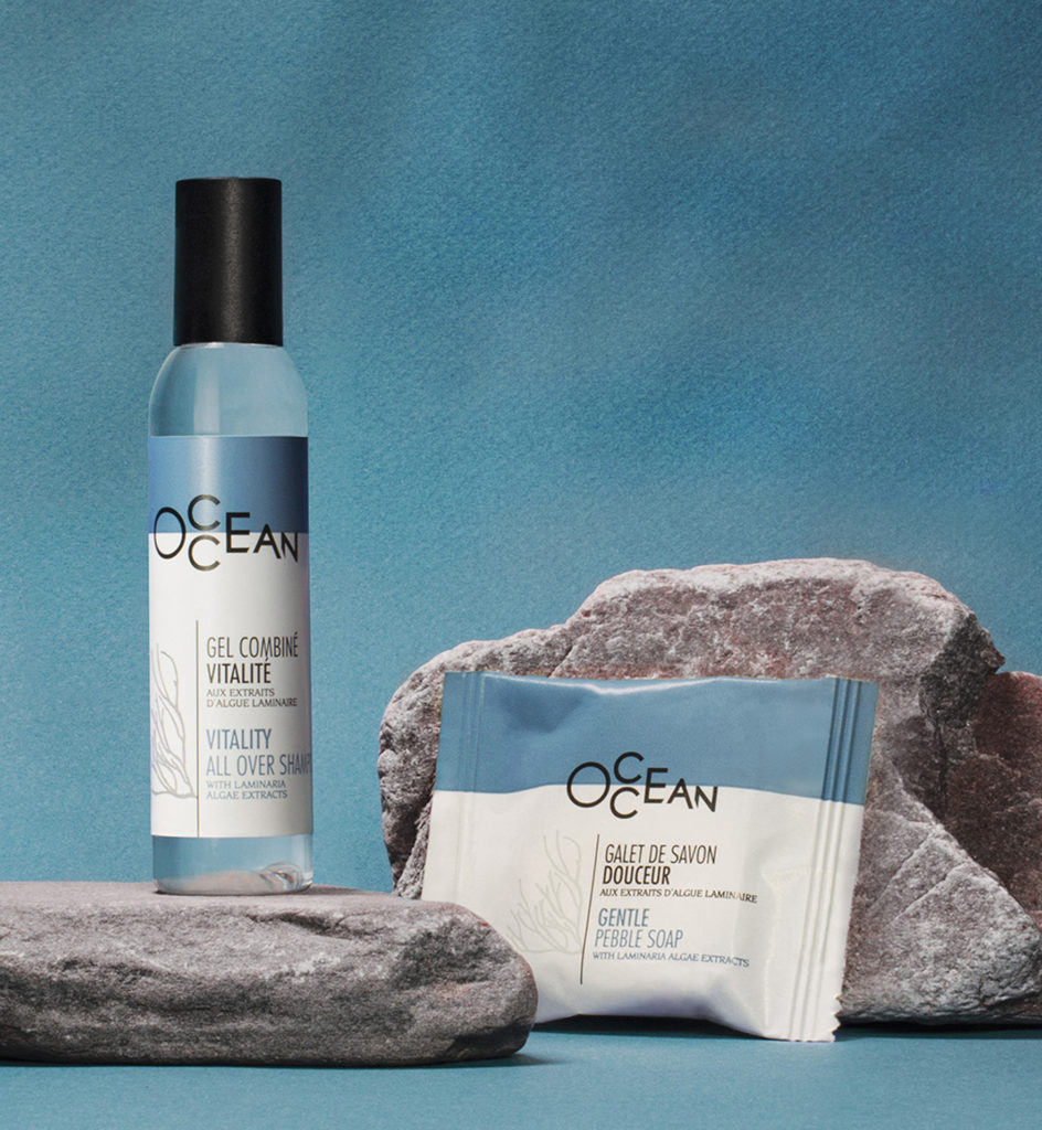 Una bottiglia di spray energizzante per il corpo accanto a una confezione di sapone delicato, entrambi esposti su rocce grigie ruvide su uno sfondo blu.