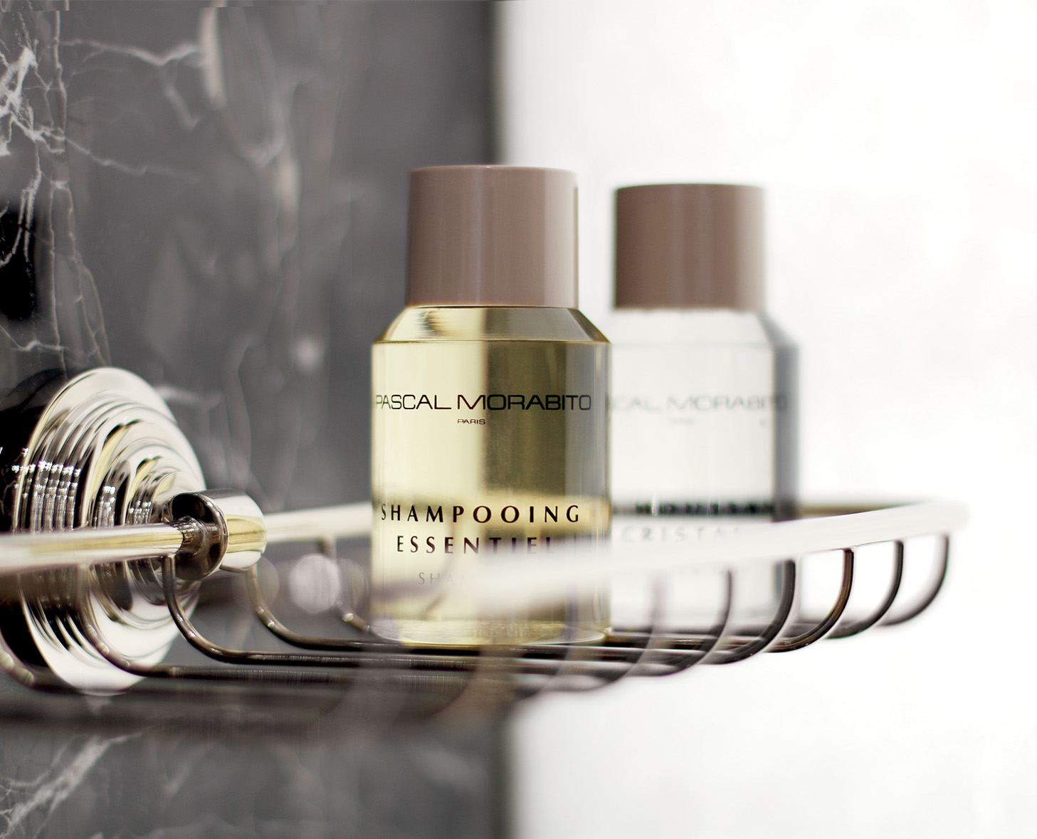 Nahaufnahme von zwei Shampooflaschen auf einem verchromten Duschkorb vor einem dunklen Marmorhintergrund. Die Flaschen sind mit eleganten Etiketten versehen, eine mit der Aufschrift „SHAMPOOING ESSENTIEL” und die andere mit „CRISTAL”.