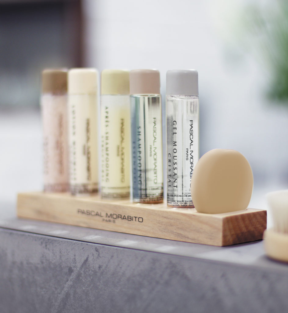 Auf einem Holzregal stehen sechs kleine Flaschen mit Hautpflegeprodukten, darunter Shampoos und Reinigungsmittel, neben einem Stück Seife. Die Flaschen sind elegant beschriftet und in einer Spa-ähnlichen Umgebung angeordnet.
