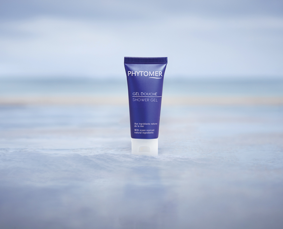 Een tube Phytomer staat op een glad, reflecterend oppervlak met een wazige strandachtergrond. De verpakking is paars en wit, wat de natuurlijke ingrediënten benadrukt.