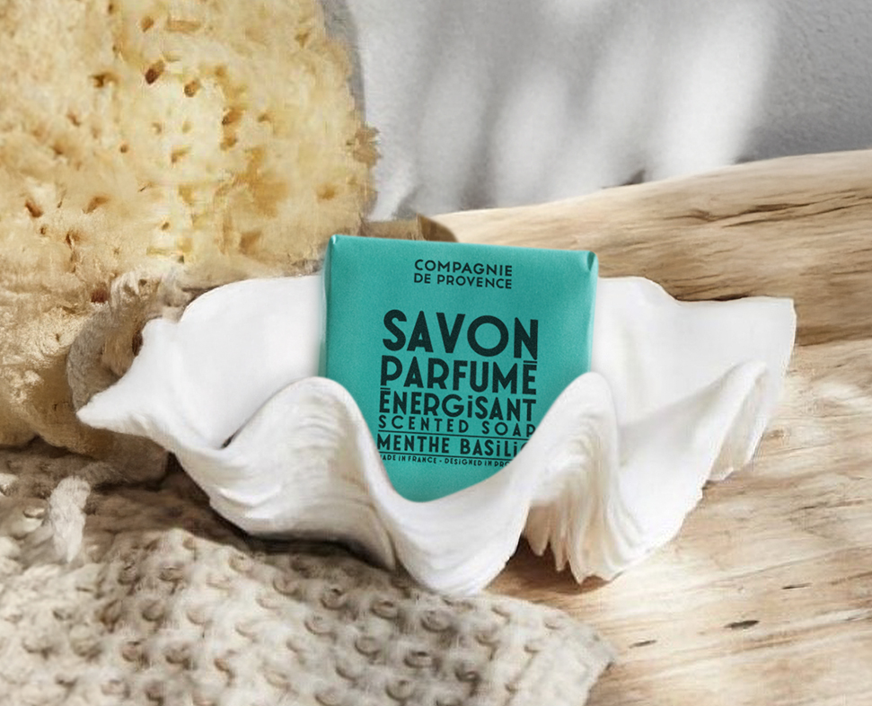 Una saponetta profumata alla menta e al basilico è elegantemente posizionata su un panno ruvido, circondata da una spugna naturale e da una superficie rustica in legno. La confezione del sapone presenta un'etichetta turchese vivace con testo in francese.