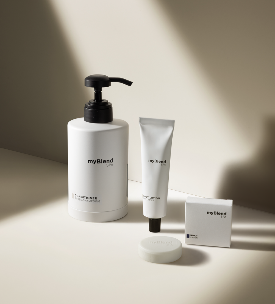 Μια μινιμαλιστική παρουσίαση των myBlend spa , που περιλαμβάνει ένα μεγάλο μπουκάλι μαλακτικό, ένα σωληνάριο λοσιόν σώματος, ένα σαπούνι και ένα μικρό κουτί, όλα τοποθετημένα σε ουδέτερο φόντο.