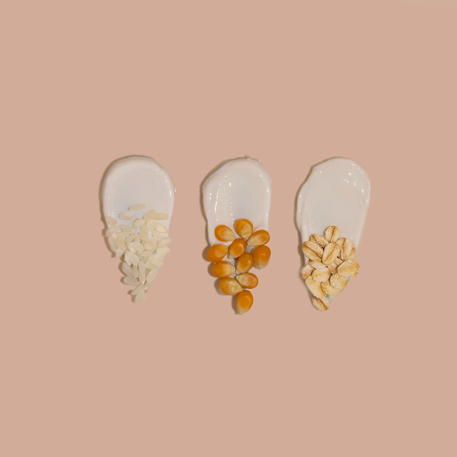 Três colheres de grãos diferentes — arroz branco, lentilhas alaranjadas e flocos de aveia — dispostas sobre um fundo rosa claro.