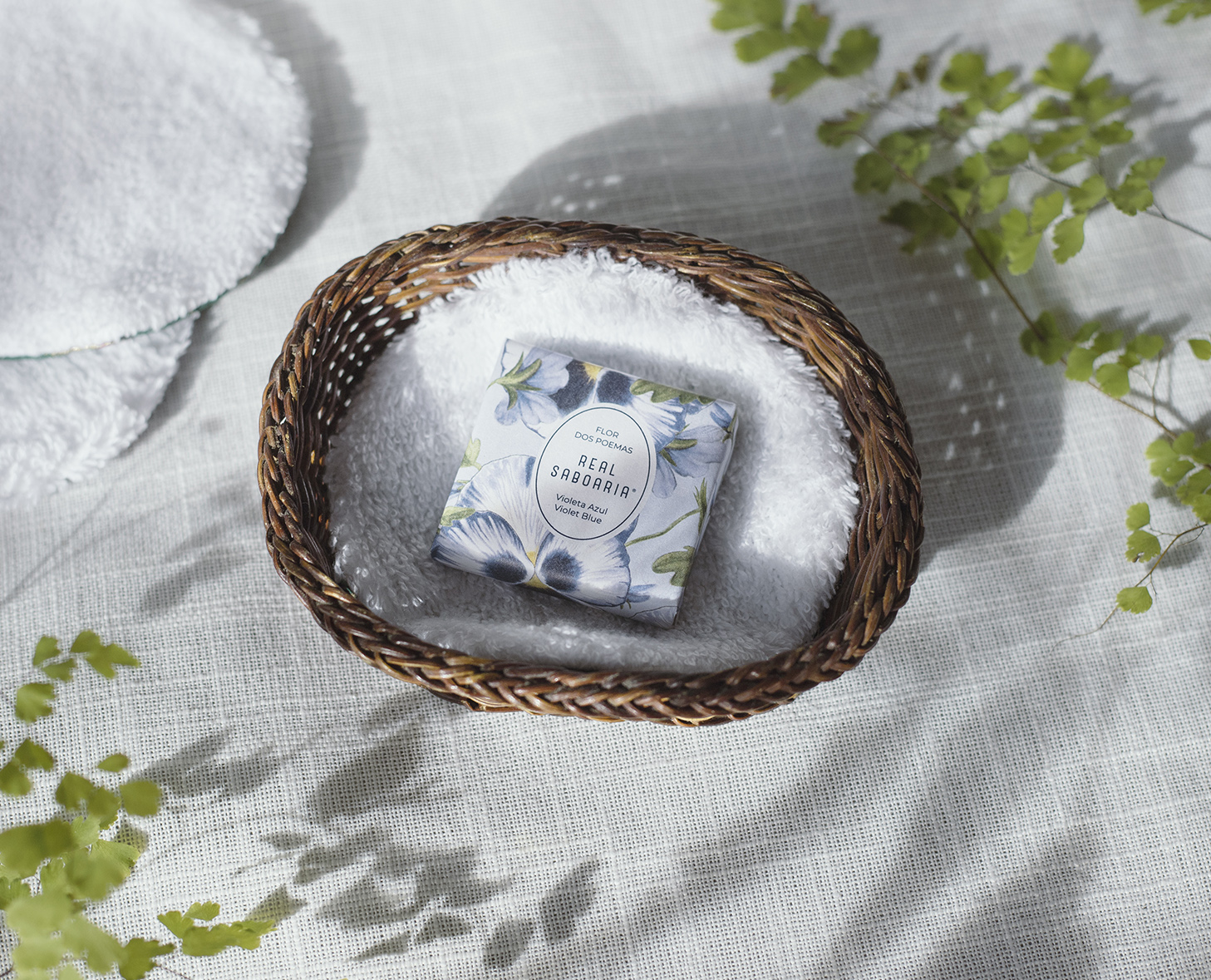 Un cestino intrecciato rivestito di tessuto bianco contiene una scatola di sapone con motivi floreali, circondata da foglie verdi su una superficie leggermente ruvida.