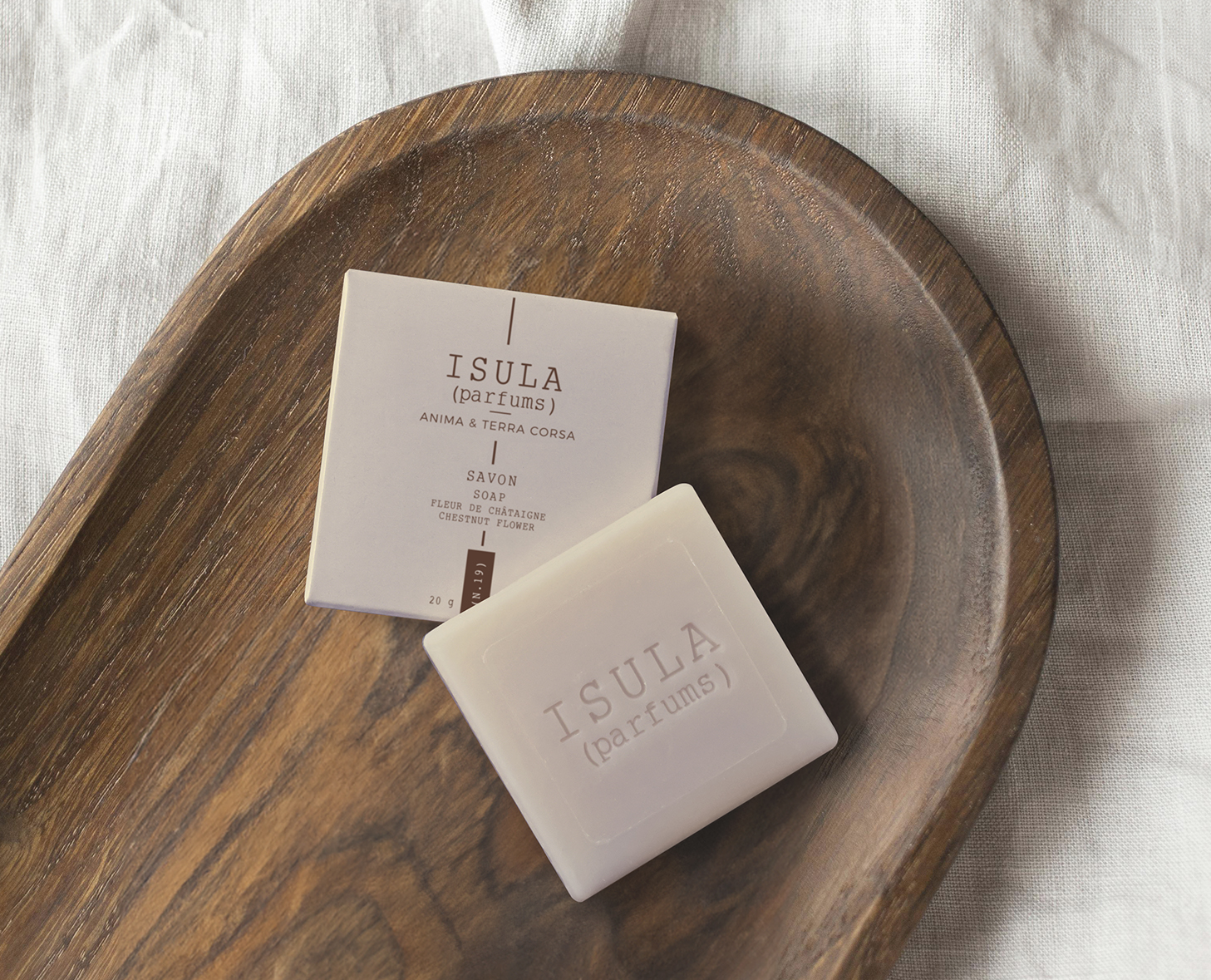 Un vassoio di legno contiene una saponetta e una confezione, entrambe contrassegnate dalla scritta "ISULA" e da una descrizione in stile minimalista. Lo sfondo è costituito da una superficie di tessuto chiaro.