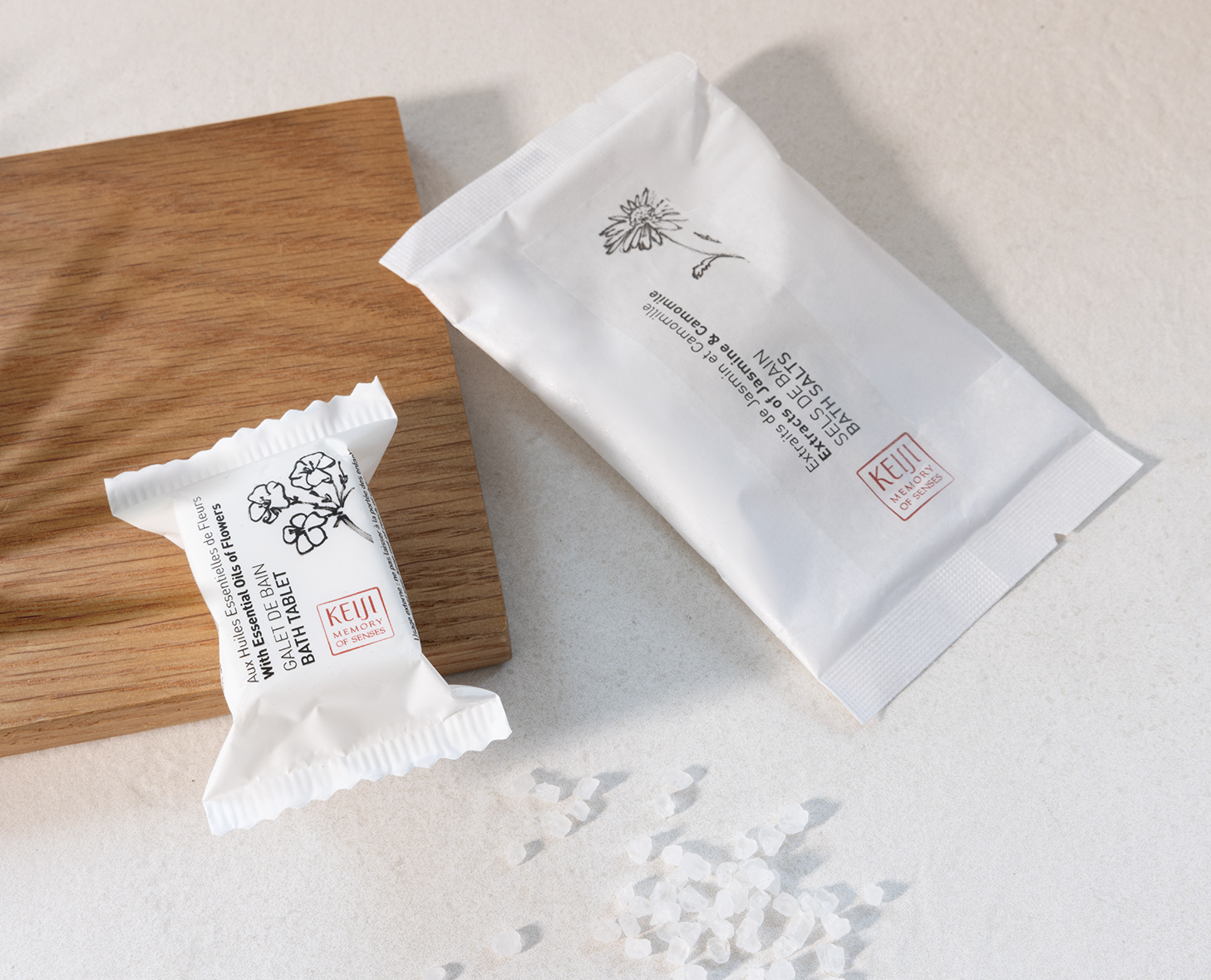 Zwei elegante Verpackungen von Hautpflegeprodukten liegen auf einem hellen Hintergrund, begleitet von einer Streuung von grobem Salz. Eine Verpackung ist in Weiß mit botanischen Illustrationen umhüllt, während die andere ein minimalistisches Design mit einer Margerite aufweist.