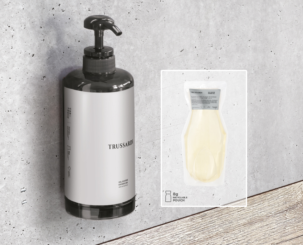 Eine schwarze Pumpflasche mit der AufschriftTrussardi” und einem eleganten Design steht vor einer strukturierten grauen Wand neben einem kleinen durchsichtigen Beutel, der eine Produktprobe enthält.