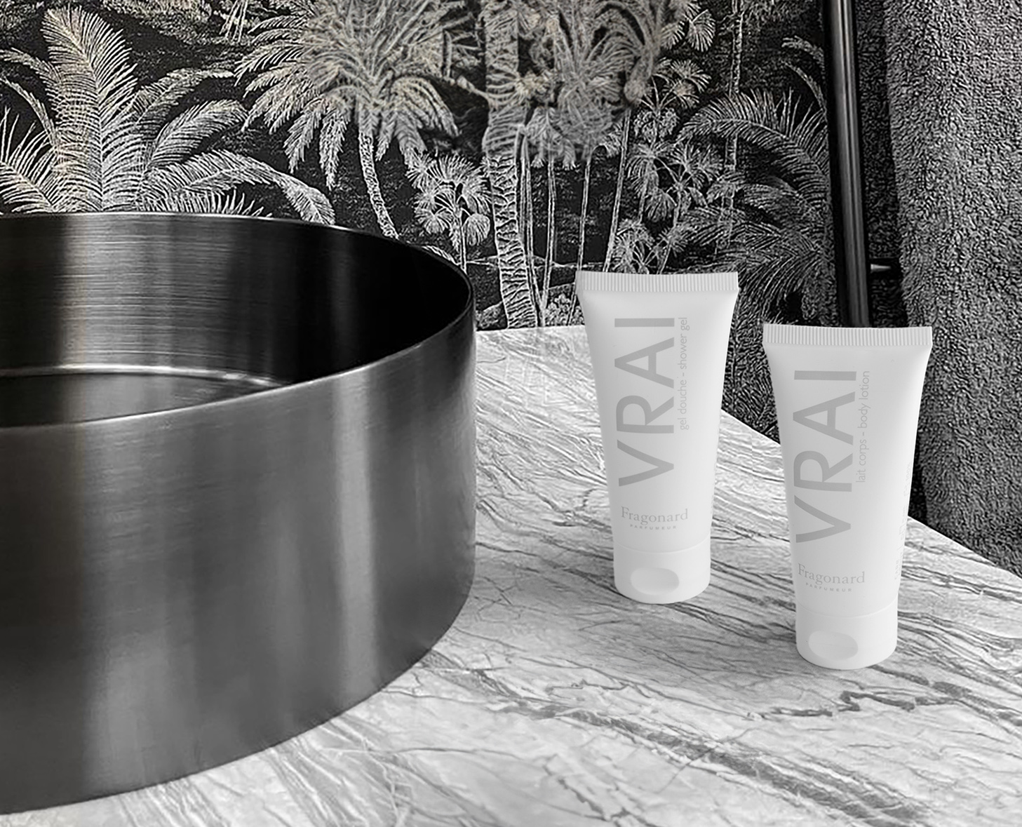 Un elegante bagno con un lavabo circolare nero e due tubi bianchi con la scritta "VRAI" su un ripiano in marmo, con una carta da parati tropicale sullo sfondo.