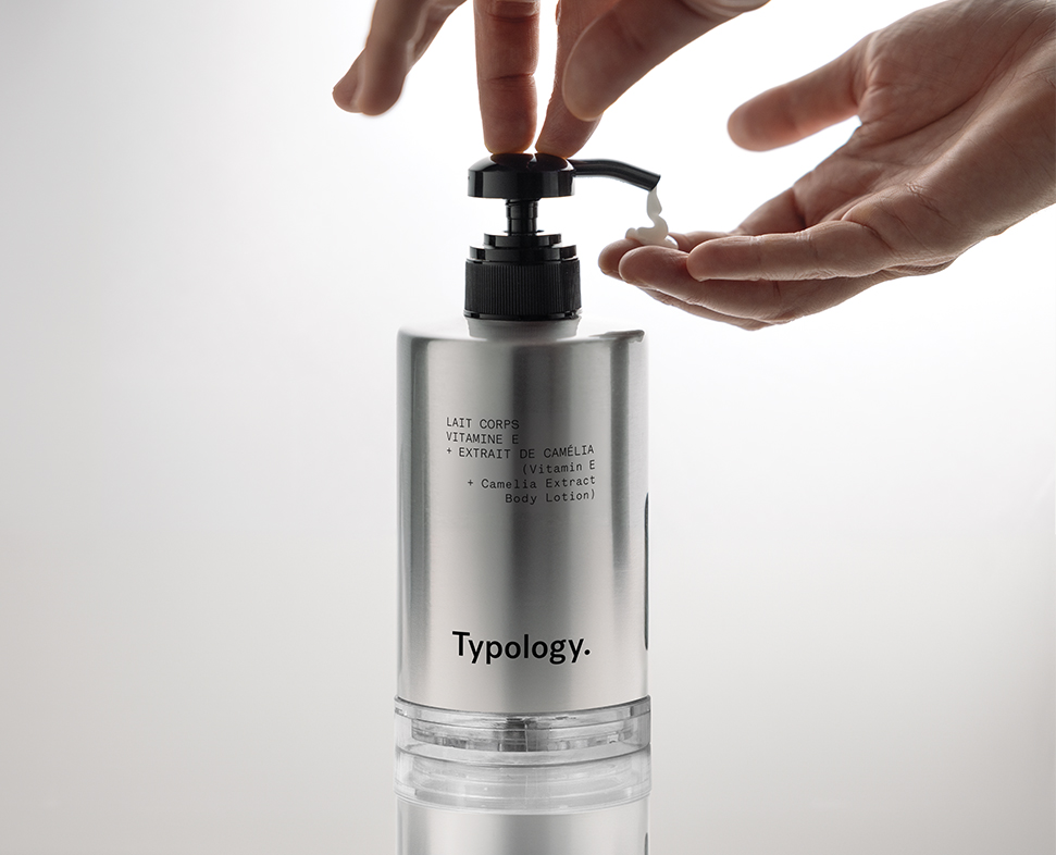 Eine Hand drückt mit einer Pumpe Körperlotion aus einer eleganten silbernen Flasche mit der AufschriftTypology und einer Liste der Inhaltsstoffe. Der Hintergrund ist sanft beleuchtet, wodurch das Produkt besonders hervorgehoben wird.