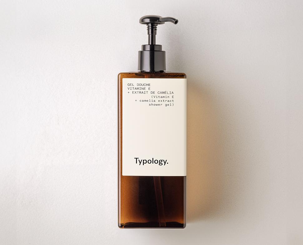 Eine braune Glasflasche mit Pumpspender für Duschgel mit der AufschriftTypology” und „Gel Douche” sowie den Hauptinhaltsstoffen Kamelienextrakt und Vitamin E. Die Flasche steht vor einem hellen Hintergrund.