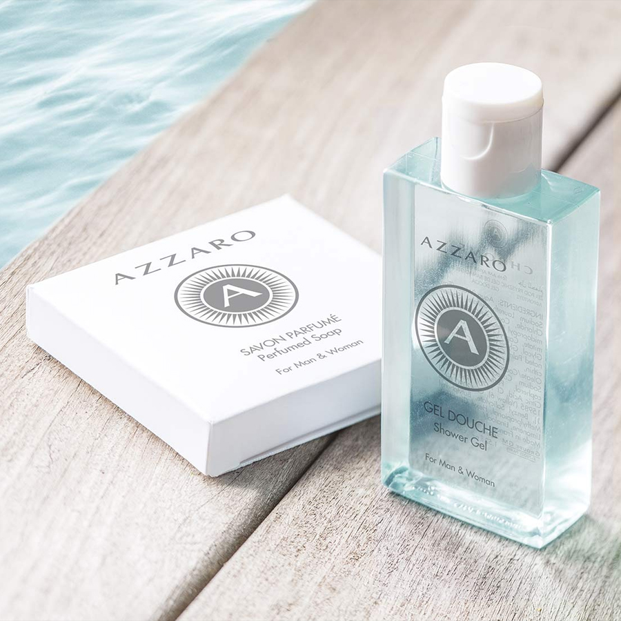 A bottle of Azzaro shower gel and a bar of Azzaro soap placed on a wooden surface beside water. Both products are labeled for men and women.