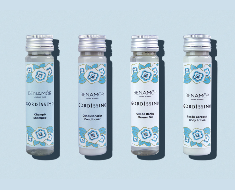 Quattro flaconi di prodotti per la cura della pelle con l'etichetta "BENAMÓR GORDÍSSIMO" e un disegno floreale blu, tra cui shampoo, balsamo, gel doccia e lozione per il corpo. I flaconi sono disposti in fila su uno sfondo azzurro.