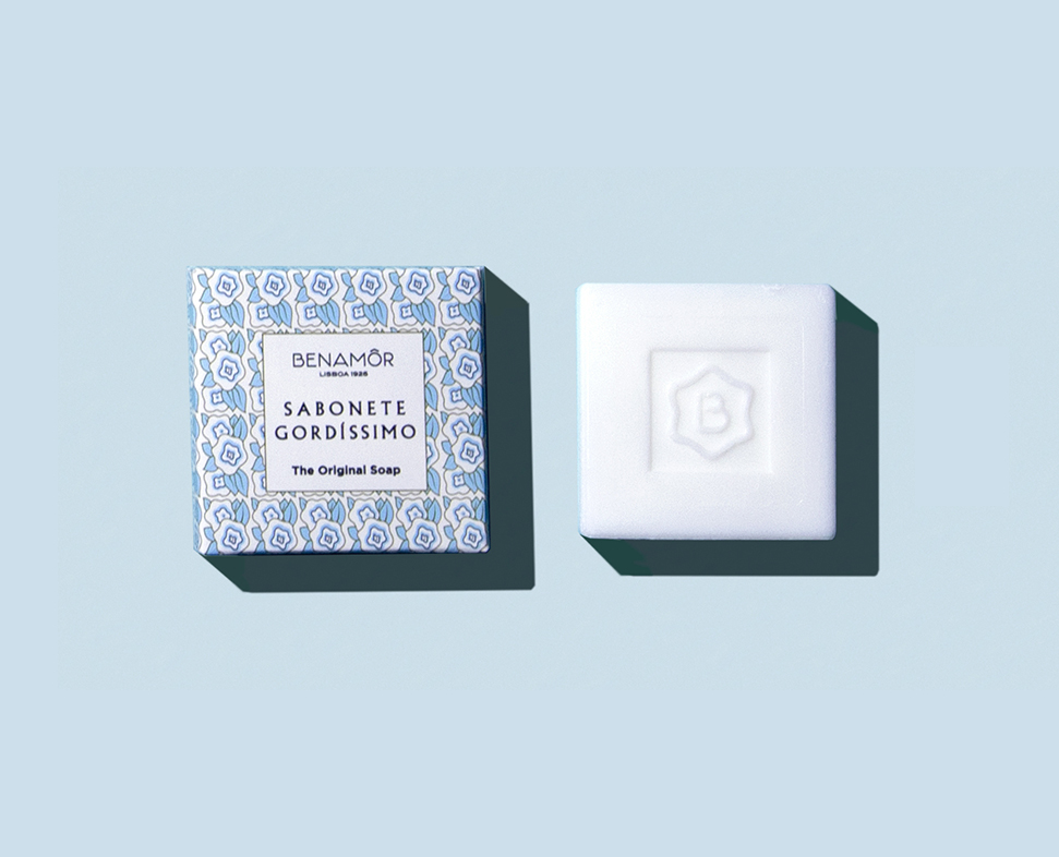 Una saponetta quadrata bianca è accanto a una scatola decorativa blu e bianca con la scritta "Benamôr Sabonete Gordíssimo - Il sapone originale". Lo sfondo è di un delicato colore blu.