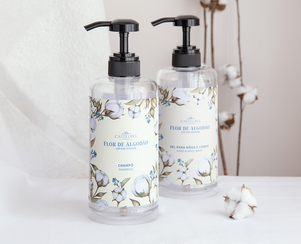 Duas garrafas transparentes de champô e gel de banho, com a marca «Flor de Algodão», com desenhos florais e tampas pretas, expostas ao lado de uma planta de algodão.