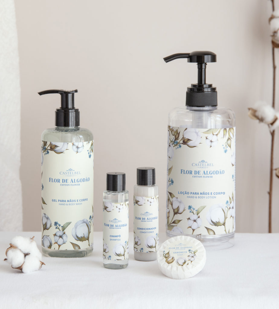 Uma coleção de produtos de higiene pessoal com embalagens florais, incluindo um gel para mãos e corpo, uma loção, um frasco pequeno e um sabonete, exibidos num fundo neutro com plantas de algodão.