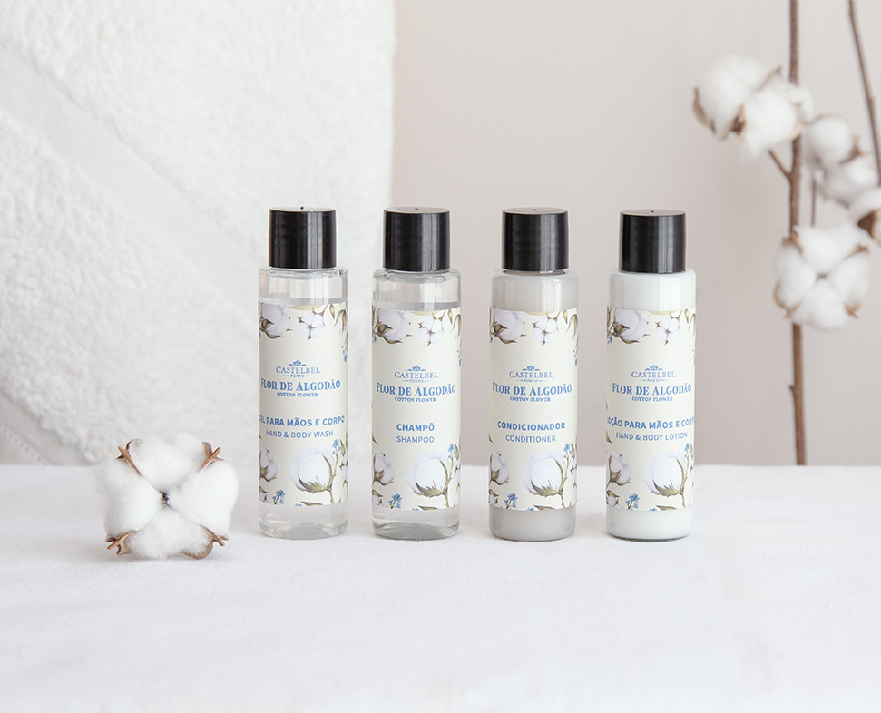 Quatro frascos de produtos para cuidados com os cabelos e o corpo, incluindo loção para as mãos, champô, condicionador e esfoliante corporal, dispostos esteticamente sobre um fundo claro, com uma planta de algodão ao lado.